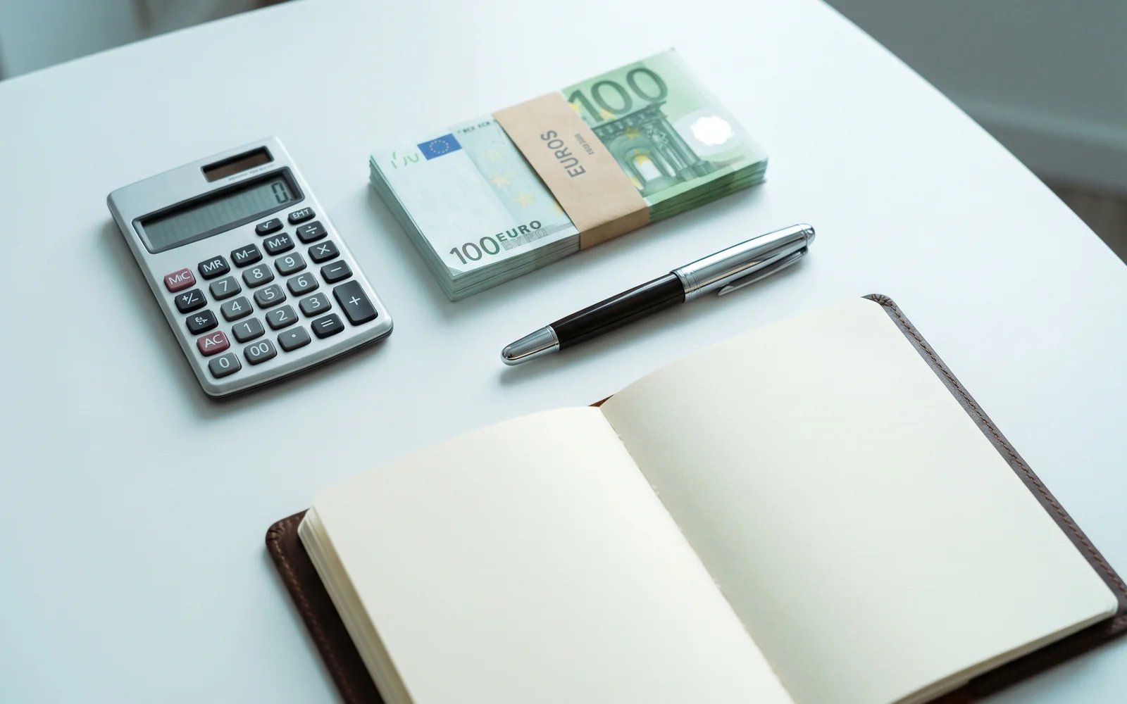 Calculatrice, liasse de billets en euros, stylo et carnet ouvert sur un bureau épuré